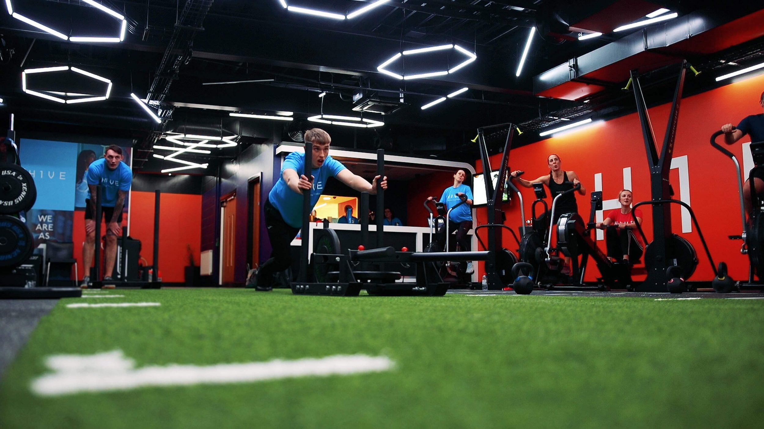 white man in a busy gym is pushing a piece of equipment down a fake grass track