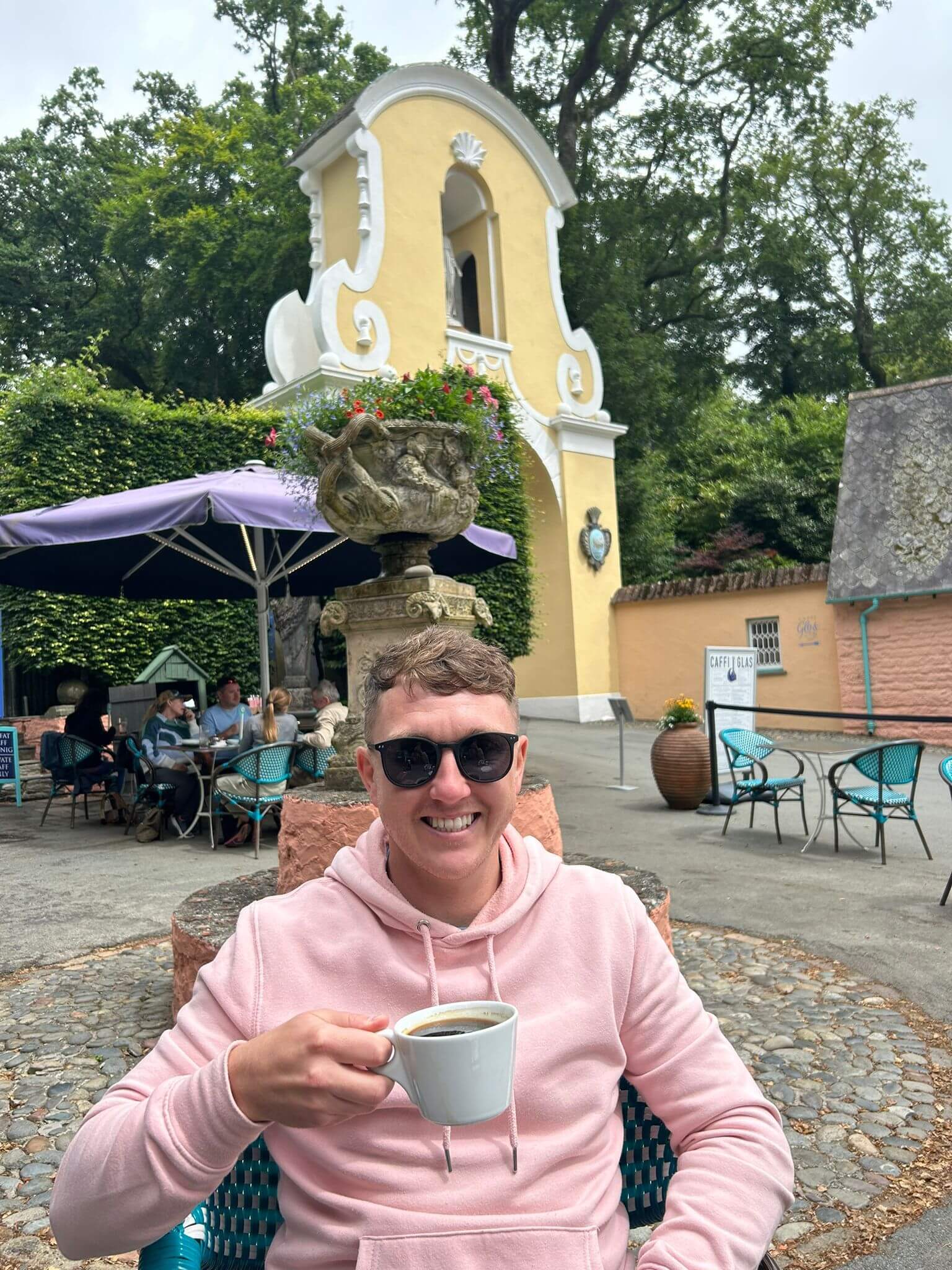 craig smiling at the camera in a pink hoodie and a cup of hot drink with his sunglasses on