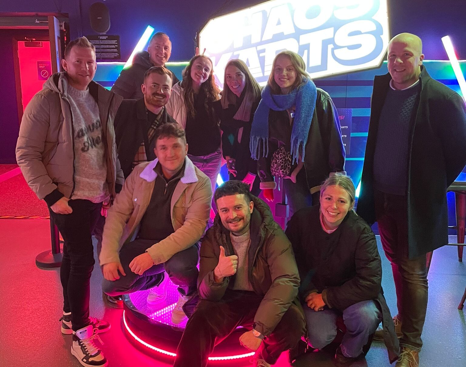 the whole team gathered around at Chaos Karts, smiling with neon lights in the background. All at varying heights.