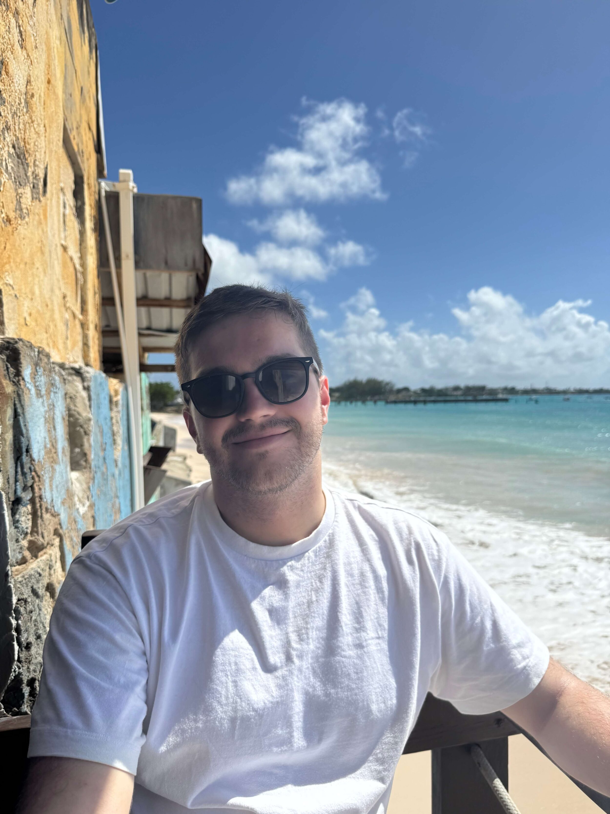 matt sat smiling at the camera on a beach with sunglasses on