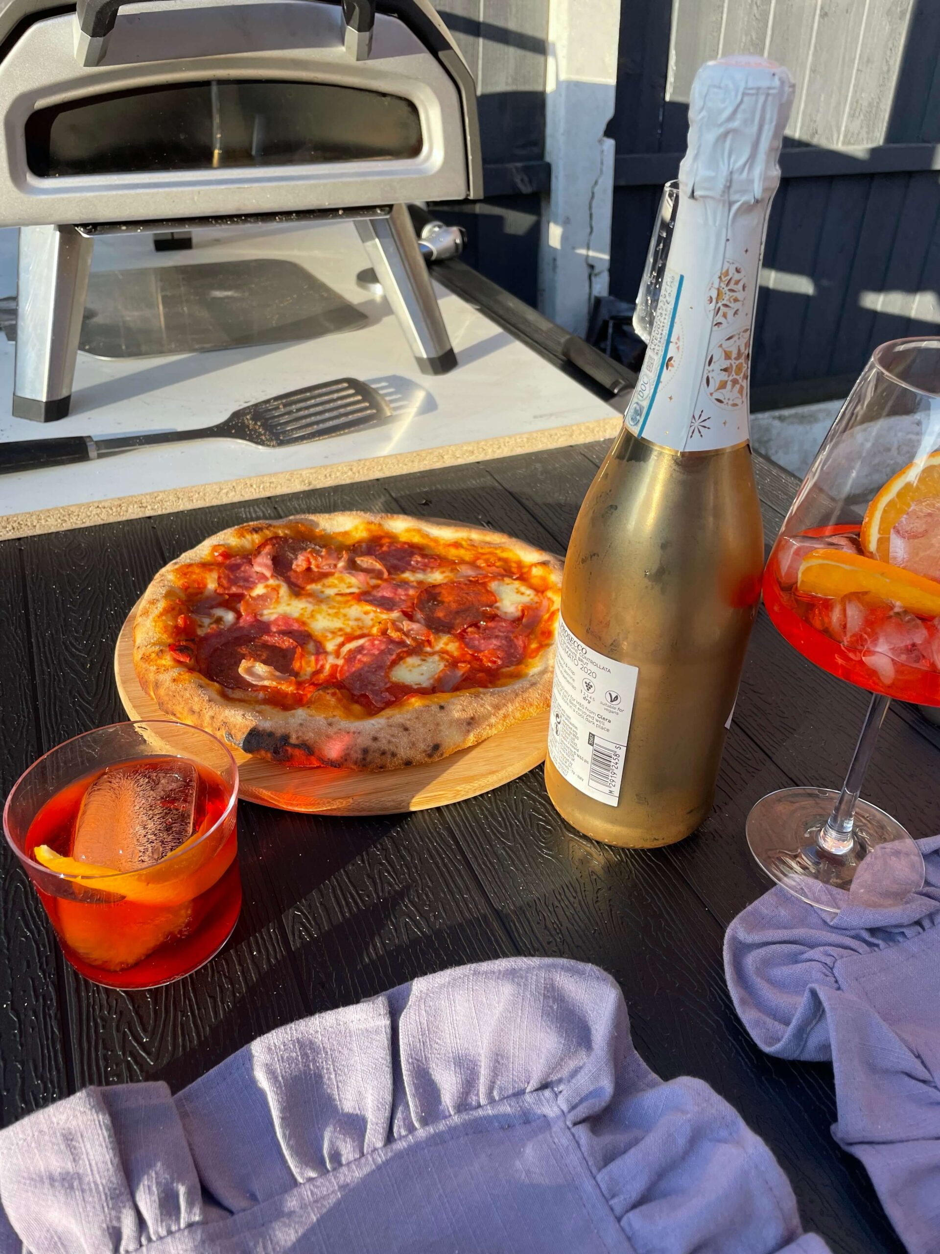 pizza oven on a tabletop with a homemade pizza, bottle of prosecco and two Aperol Spritz.